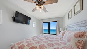 A bedroom with a bed, a ceiling fan, a TV, a window with a view, and a door.
