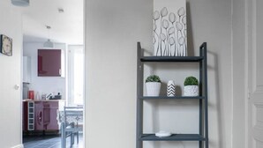Un interior moderno con piso de madera, un estante metálico gris con objetos decorativos, y vista a una zona de cocina.
