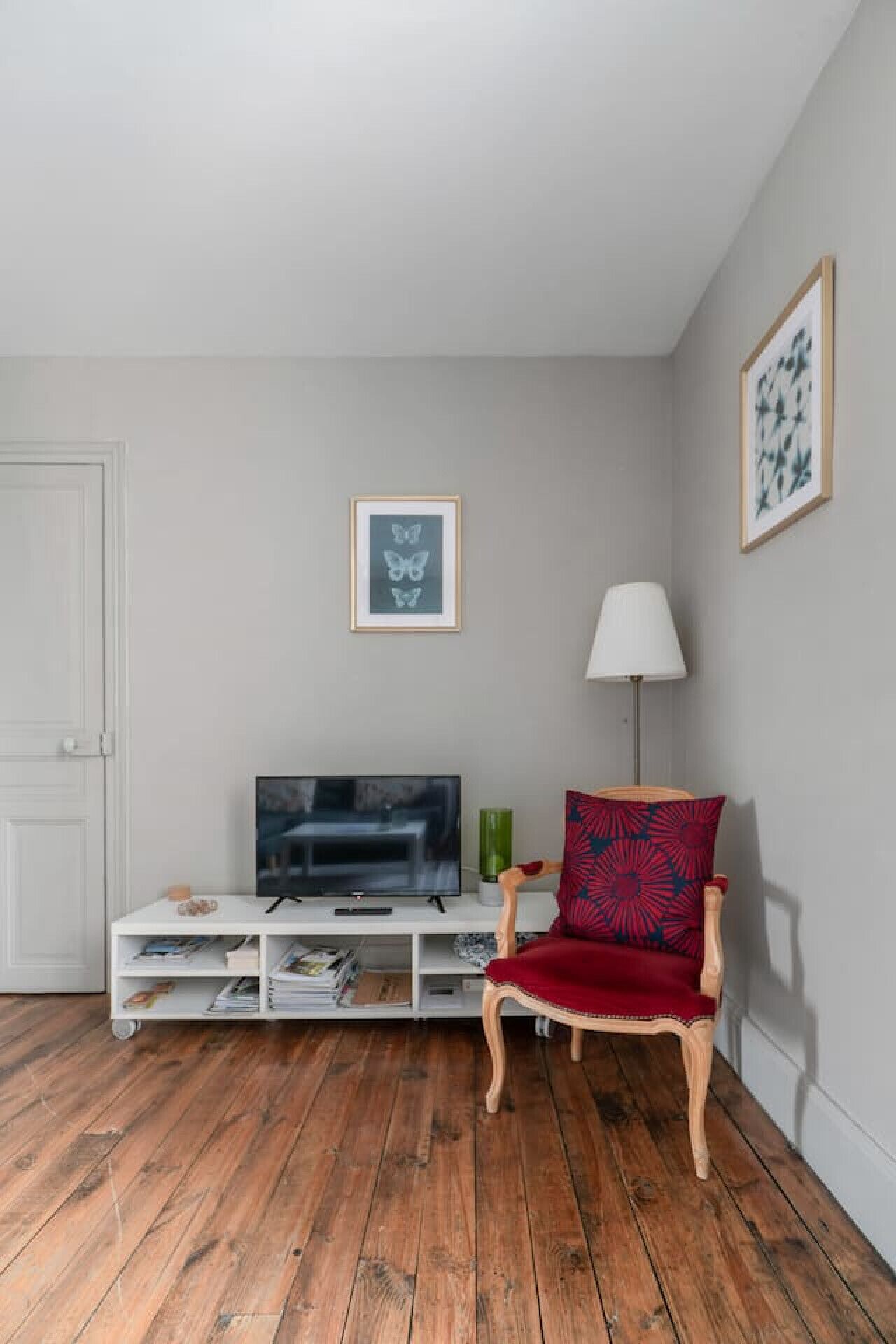 Ein Wohnzimmer mit Fernseher auf einer weißen Unterhaltungsecke, einem roten Sessel, einer Stehlampe und zwei gerahmten Bildern an der Wand.