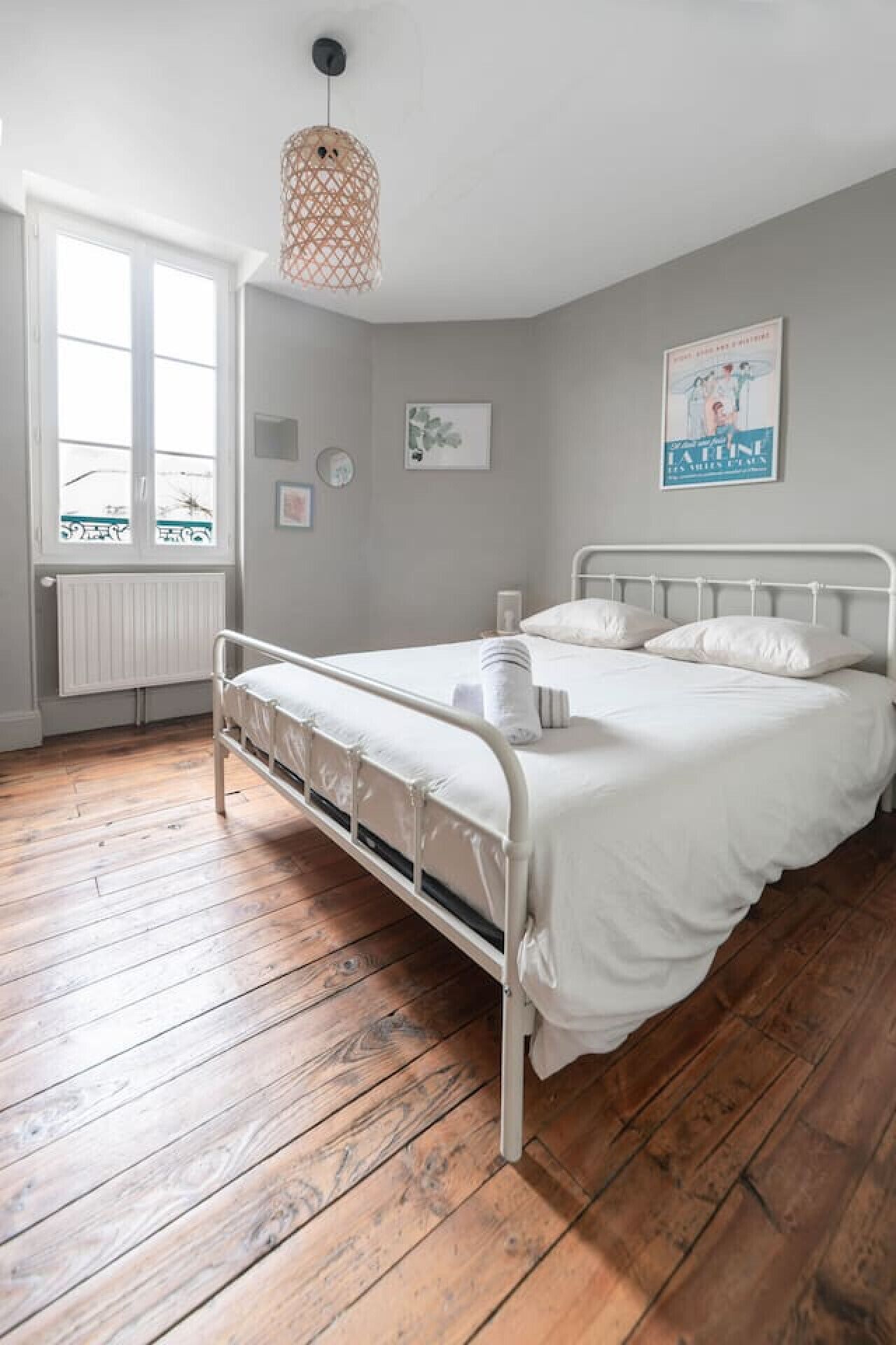Een slaapkamer met een bed van wit metaal, een houten vloer, een witte radiator, een raam en een hanglamp.