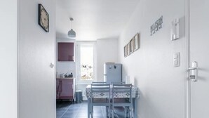 Un pasillo estrecho con suelo de madera que conduce a un comedor con mesa y sillas. Un reloj se ve en la pared izquierda, y hay un refrigerador al fondo.