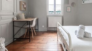 Un dormitorio con suelo de madera, una cama con ropa de cama blanca, un escritorio con una silla, una lámpara colgante y una ventana con vistas.