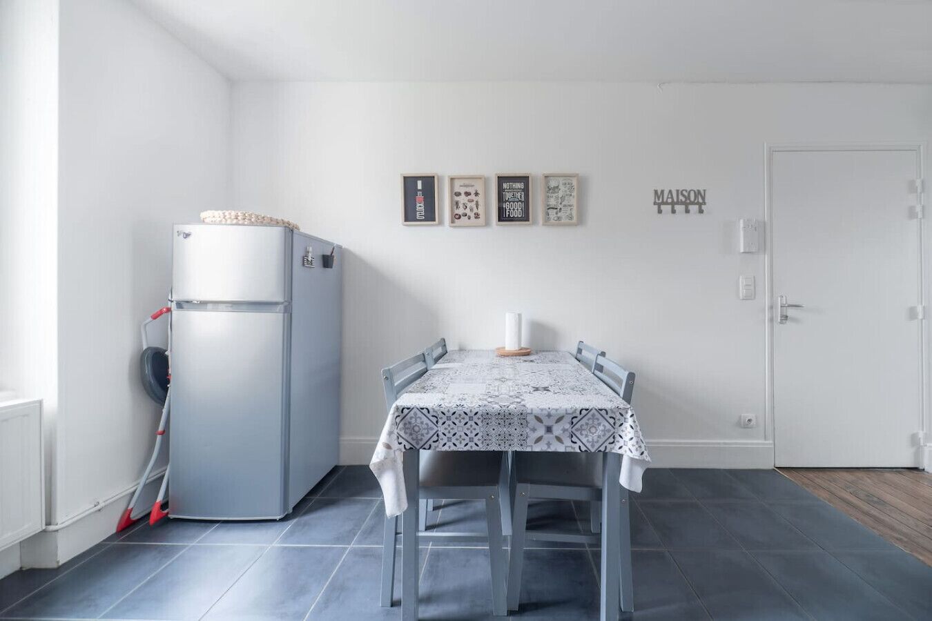 Une petite cuisine moderne avec un réfrigérateur gris, une table à manger recouverte d’une nappe à motifs et une porte blanche.