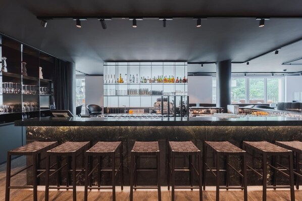 A modern bar area with a long counter, multiple stools, and shelves stocked with bottles and glasses.