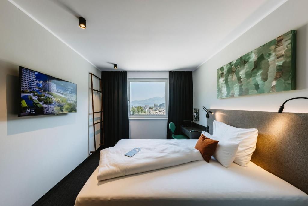 A hotel room with a bed, a TV mounted on the wall, a window with a view, and a painting on the wall.