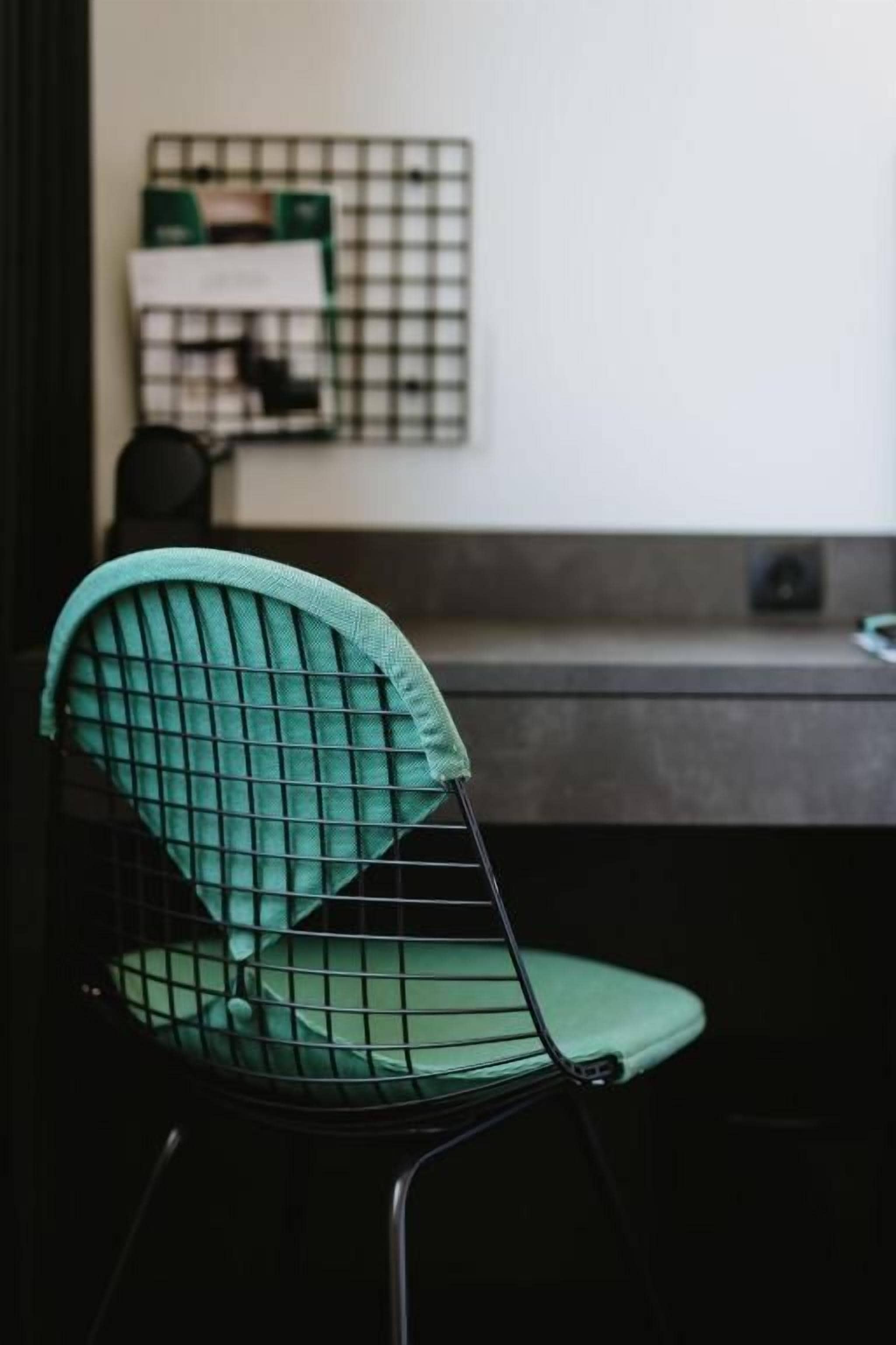 A chair with a wireframe backrest and a cushion, placed in front of a desk with a grid-patterned wall behind it.