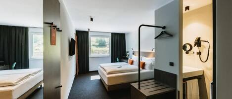 A hotel room with two beds, a desk, a bathroom with a mirror and towel rack, and a view of the outdoors.