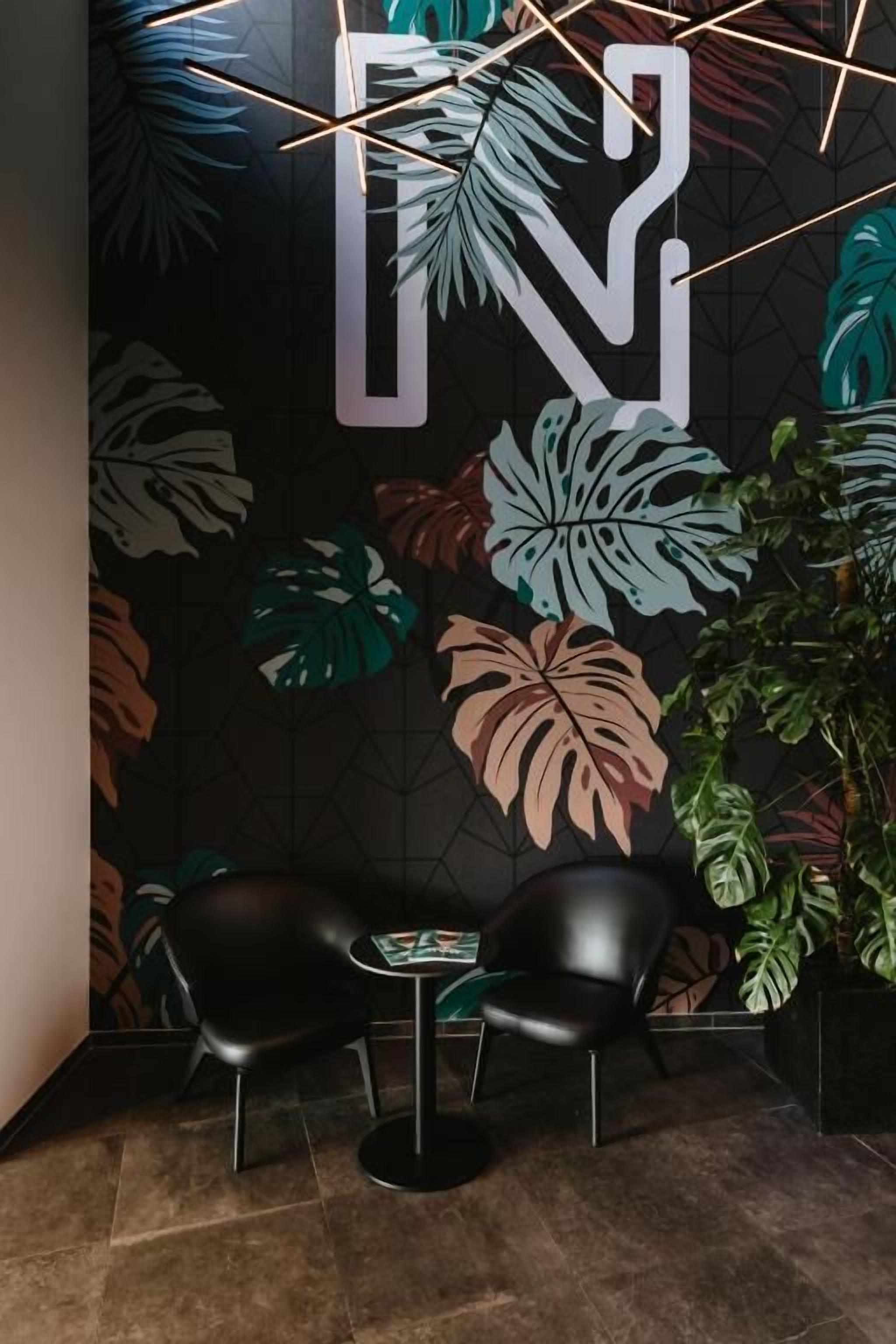 Zona de recepción con un gran mural en la pared, dos sillas negras, una mesita y una planta en maceta.