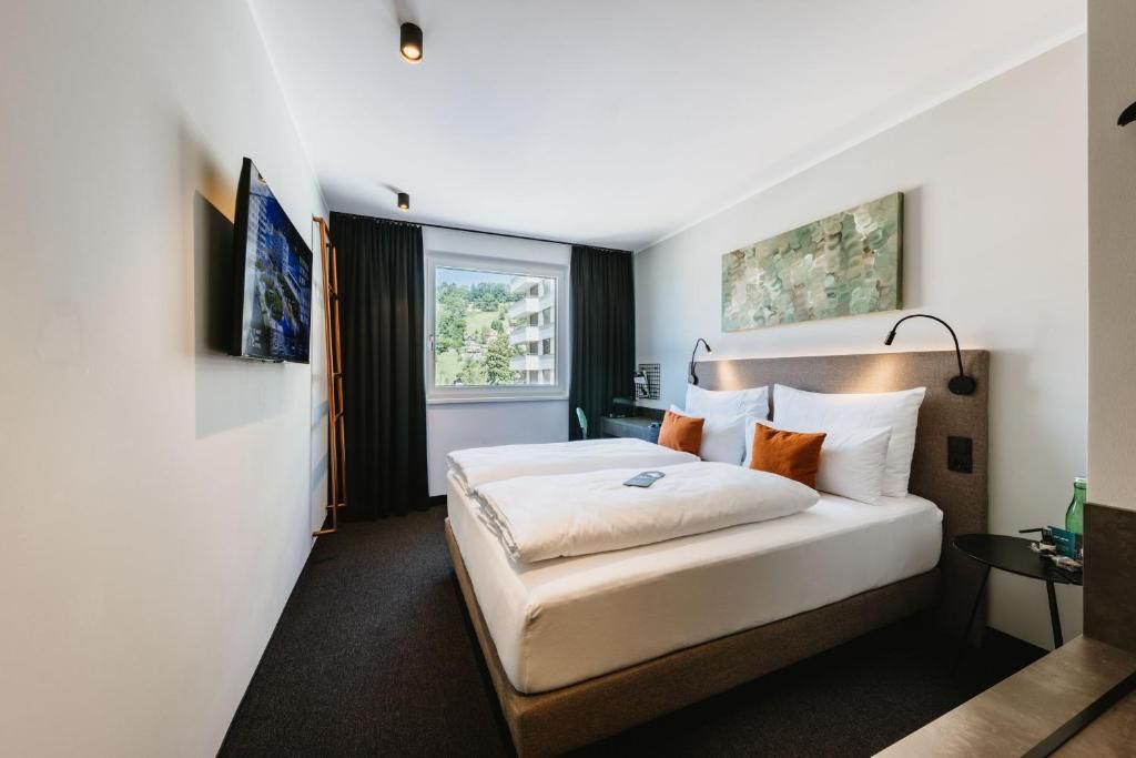 A hotel room with a bed, a nightstand, a lamp, a window with curtains, and artwork on the walls.