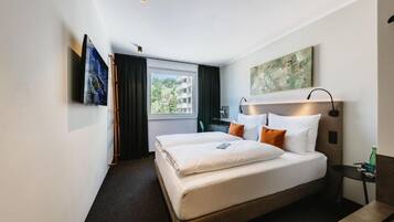 A hotel room with a bed, a nightstand, a lamp, a window with curtains, and artwork on the walls.
