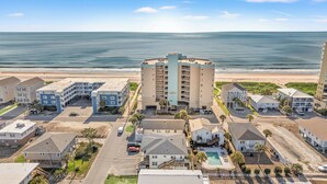 A coastal hotel complex with multiple buildings, including a prominent tower with balconies, a parking area, and a swimming pool. - Hyperion 604 Oceanfront Sleeps 8-Outdoor Pool (North Myrtle Beach)