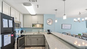 A modern kitchen with grey cabinets, a black refrigerator, a microwave, and a sink. There is a clock on the wall and a framed quote above the counter. - Hyperion 604 Oceanfront Sleeps 8-Outdoor Pool (North Myrtle Beach)