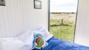 A bedroom with a bed, white walls, a window with a view of a grassy field, and a framed picture on the wall. - Tiny Balloon House by Tiny Away (Canowindra)