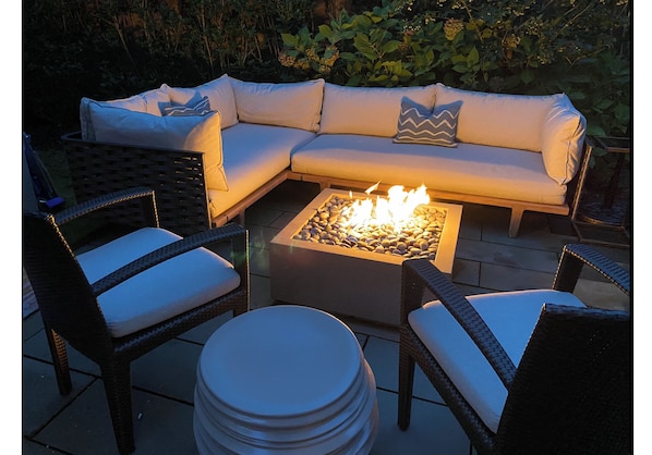 A sectional sofa with white cushions and a zigzag pillow, surrounded by wicker chairs with white cushions. In front of the sofa is a square fire pit with rocks and flames.