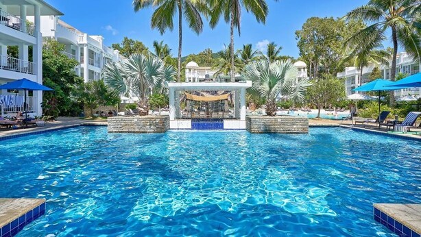 A swimming pool with a central water feature, surrounded by palm trees and lounge chairs.