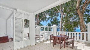A covered patio with a wooden table and chairs, white railings, and a view of palm trees and the sea.