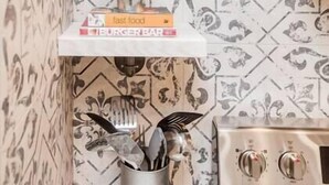 A kitchen with patterned wallpaper, a white shelf with a plant, a book titled 'Burger Bar', and a utensil holder with a spoon. - OSU 6D · Newly Renovated (6D) OSU/Central ❤Near EVERYTHING (Columbus)