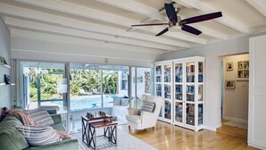 A spacious living room with a large window, a ceiling fan, a pool visible outside, and a bookshelf filled with books. - Tropical Paradise, walking distance to the beach and heated pool! (Hollywood)