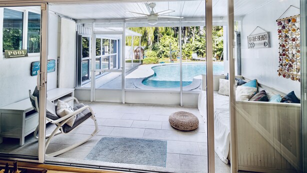 A room with a glass door leading to a pool area, a bed with pillows, a small round seating area, and a ceiling fan. - Tropical Paradise, walking distance to the beach and heated pool! (Hollywood)