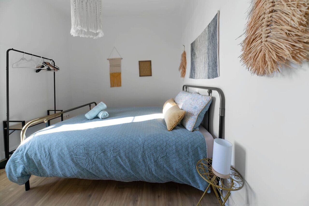 A bedroom with a bed, a hanging light, a wall-mounted macrame, and a small table with a lamp.