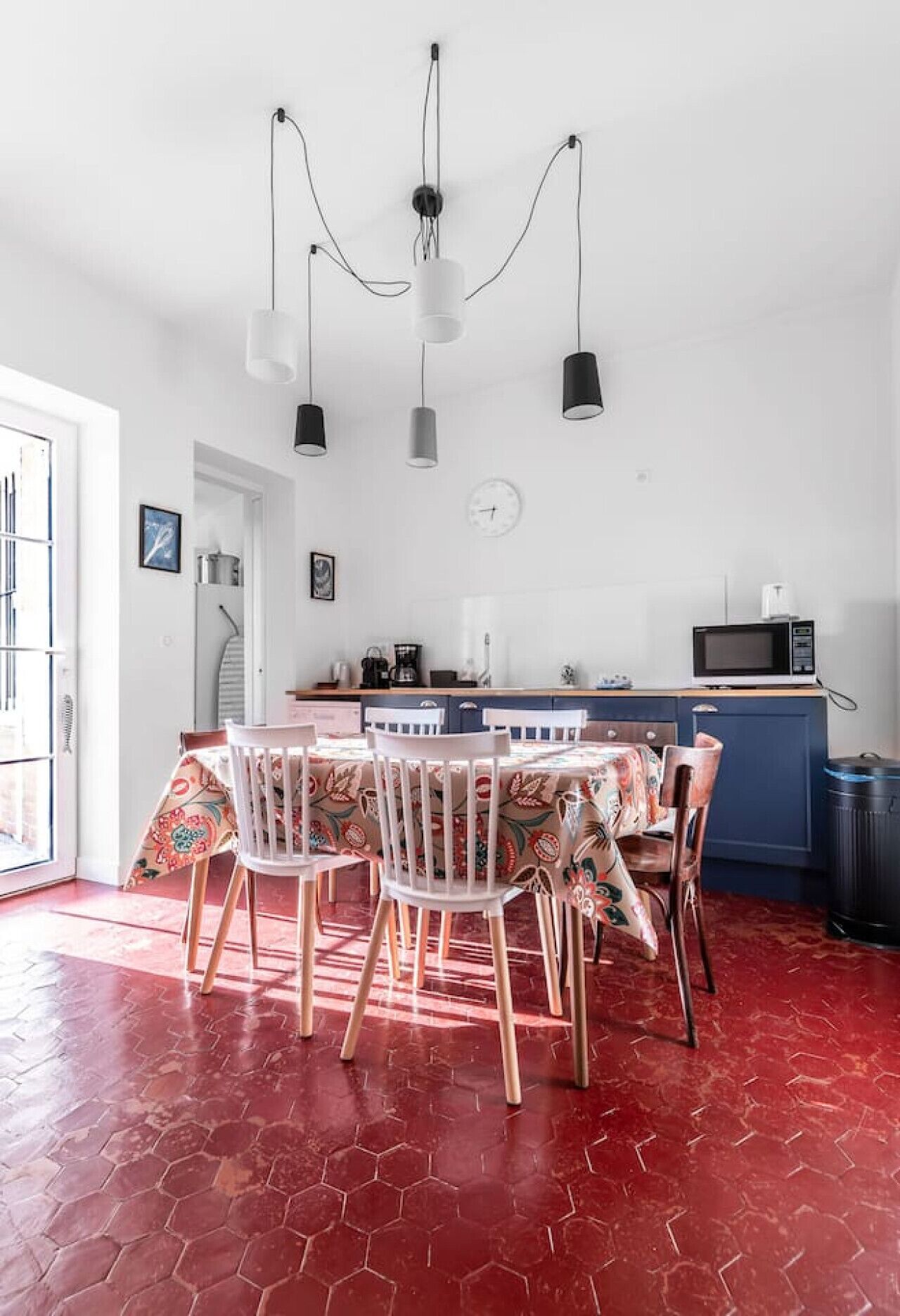 Un espace repas avec un sol hexagonal rouge, des chaises blanches, une table recouverte d’une nappe à motifs floraux et des luminaires suspendus au-dessus.