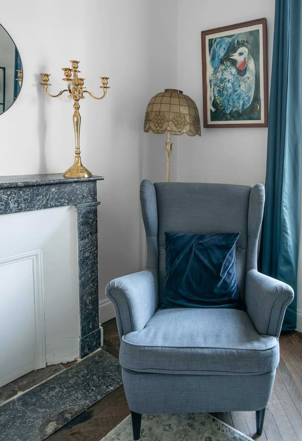A room with a grey armchair, a gold candelabra, a floor lamp, and a framed painting on the wall.