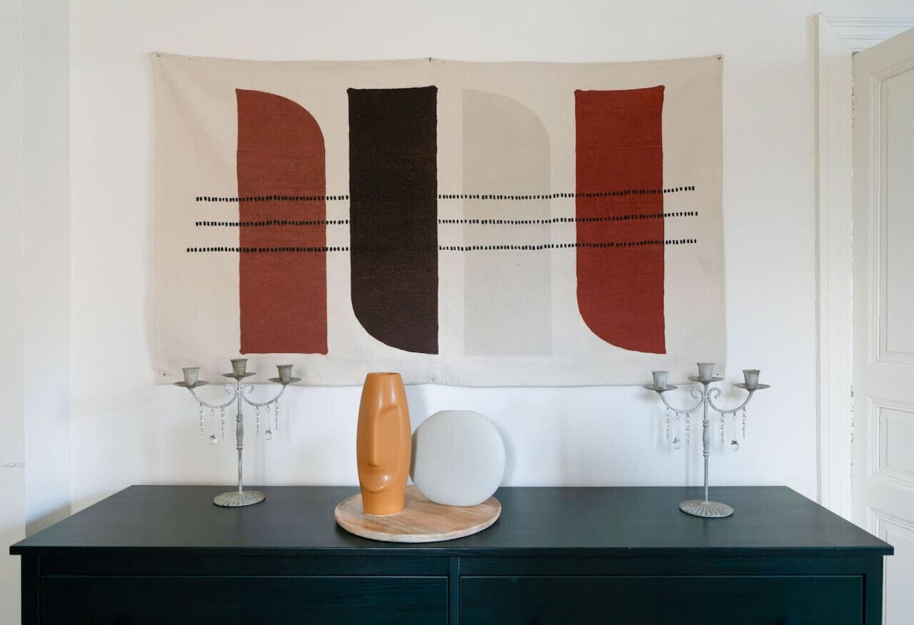 A room with a dark green dresser, a tapestry with abstract shapes, and a vase on the dresser.