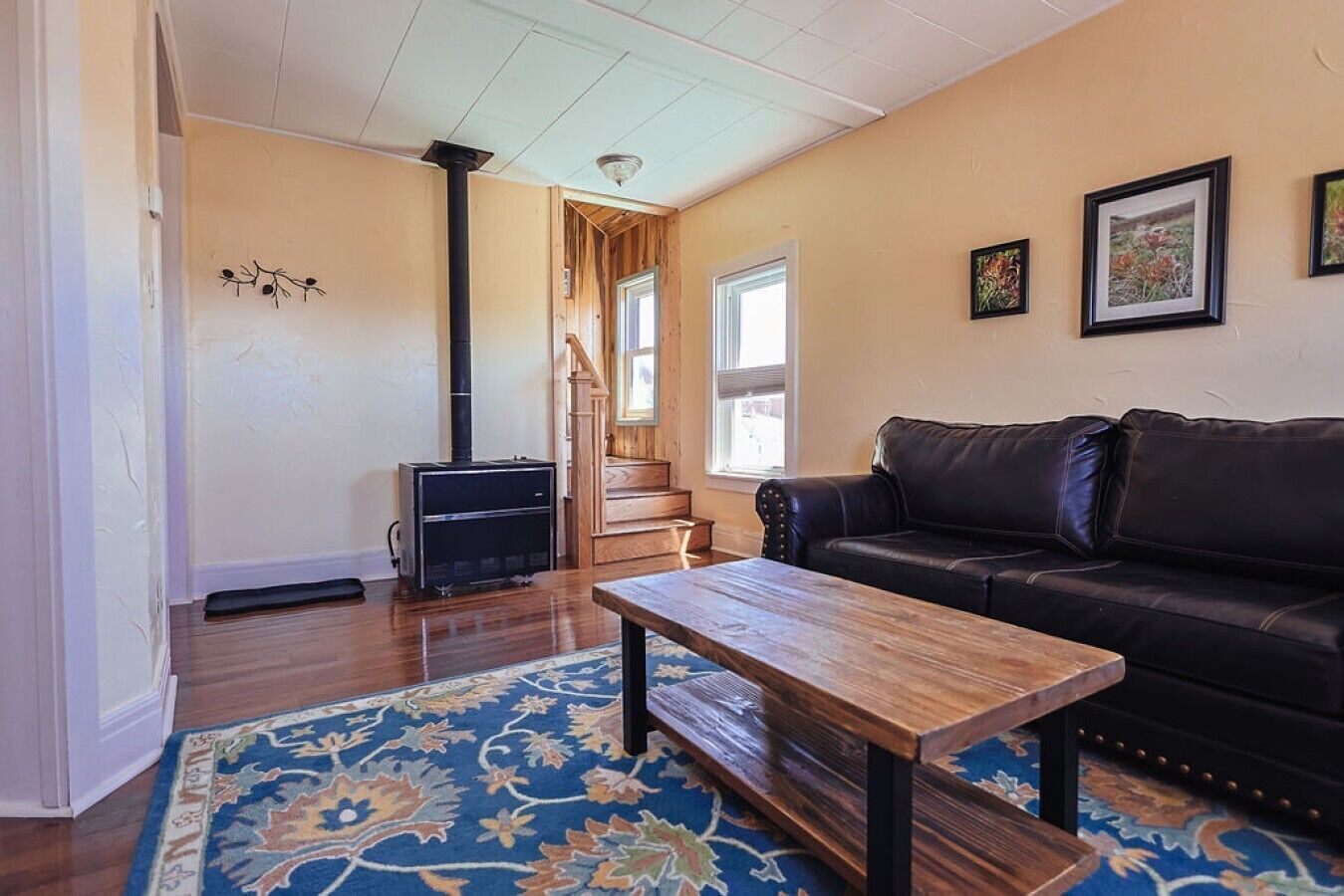 A living room with a black leather sofa, a wooden coffee table, a patterned rug, a wood-burning stove, and a staircase leading to another room.