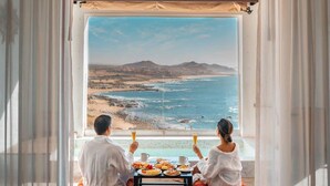 Una habitación con un amplio ventanal que da a una playa y el océano, dos personas sentadas a una mesa con desayuno, y vistas a colinas distantes.