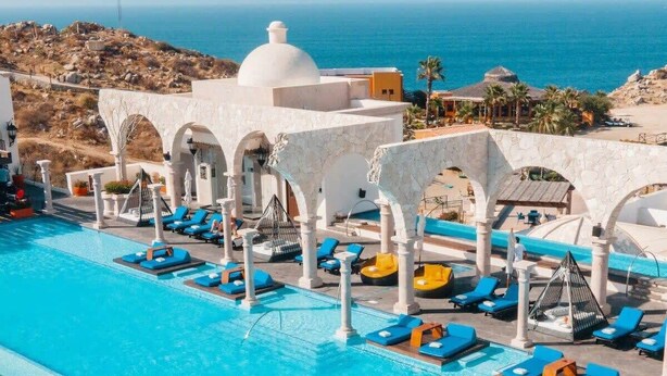 Una amplia piscina al aire libre con agua azul, rodeada de columnas y arcos blancos, y vista al océano al fondo.
