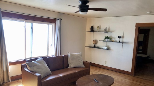 Un soggiorno con un divano in pelle marrone, un tavolino rotondo e una ventola a soffitto. Sono presenti scaffali con oggetti decorativi e una finestra con tende.