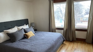Un letto rifatto con ordine, coperto da una trapunta grigia e decorato con molti cuscini, un pavimento in legno e una finestra con tende.