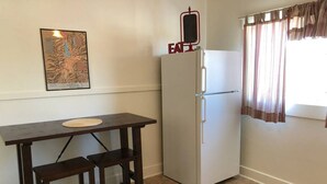 Una cucina compatta con un frigorifero bianco, un tavolo di legno con due sgabelli, un quadro appeso alla parete e una finestra con tende.