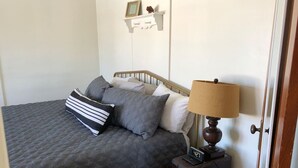 Una camera da letto con un letto, cuscini, un comodino con una lampada, una sveglia e una mensola a muro con una cornice.