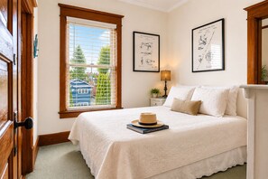 A bedroom with a bed, a window with blinds, a lamp, and two framed pictures on the wall.