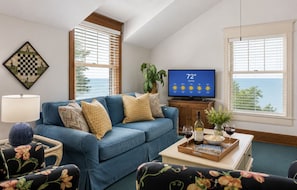 Una sala de estar con un sofá azul, un televisor mostrando un pronóstico de tiempo soleado y vista al mar a través de amplios ventanales.