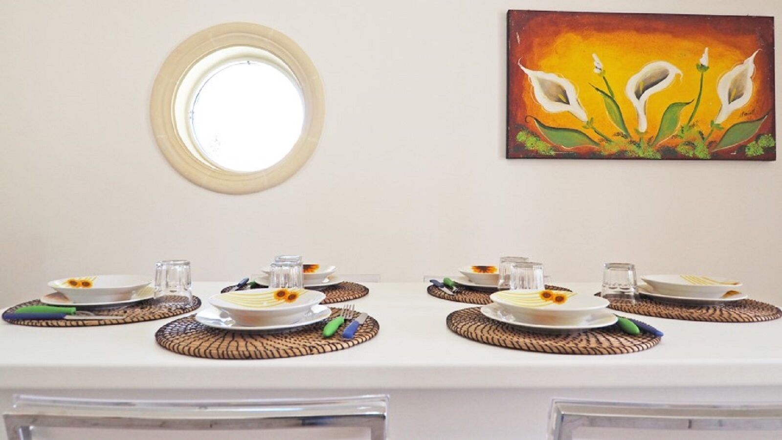Una mesa de comedor dispuesta con platos, vasos y cubiertos, con un cuadro y un espejo redondo encima.