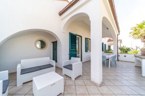 Ein überdachter Außenbereich mit weißen Korbmöbeln, grünen Fensterläden und einem Fliesenboden.