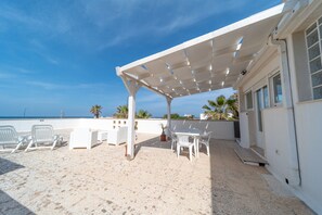 Ein überdachter Außenbereich mit weißen Möbeln, einer Pergola und Blick auf das Meer.