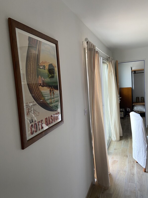 A hallway with a framed poster, a door with a curtain, and a bed with a white cover.