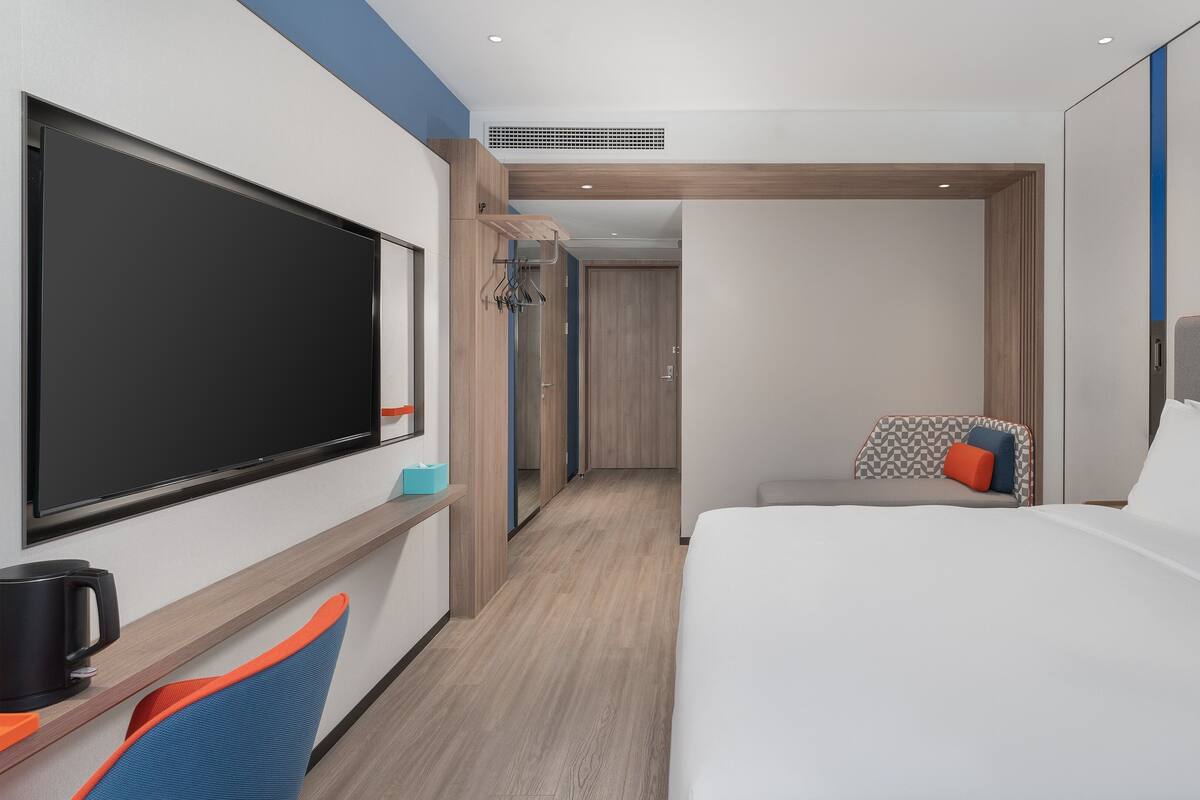 A modern hotel room with a large flat-screen TV, a bed with white linens, a seating area with a chair and cushions, and a wooden floor.