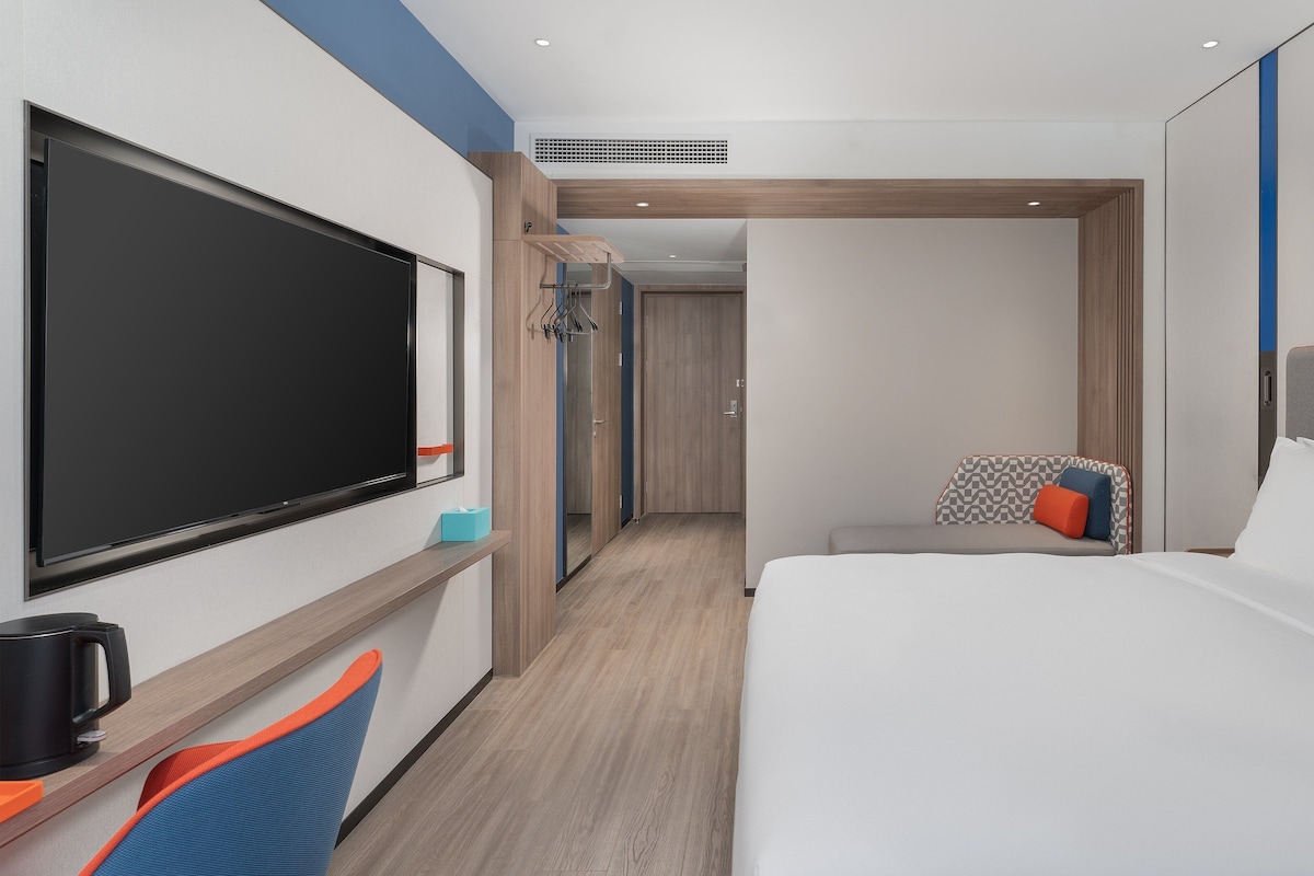 A hotel room with a large flat-screen TV, a bed with white bedding, a seating area with a chair, and a wooden floor.