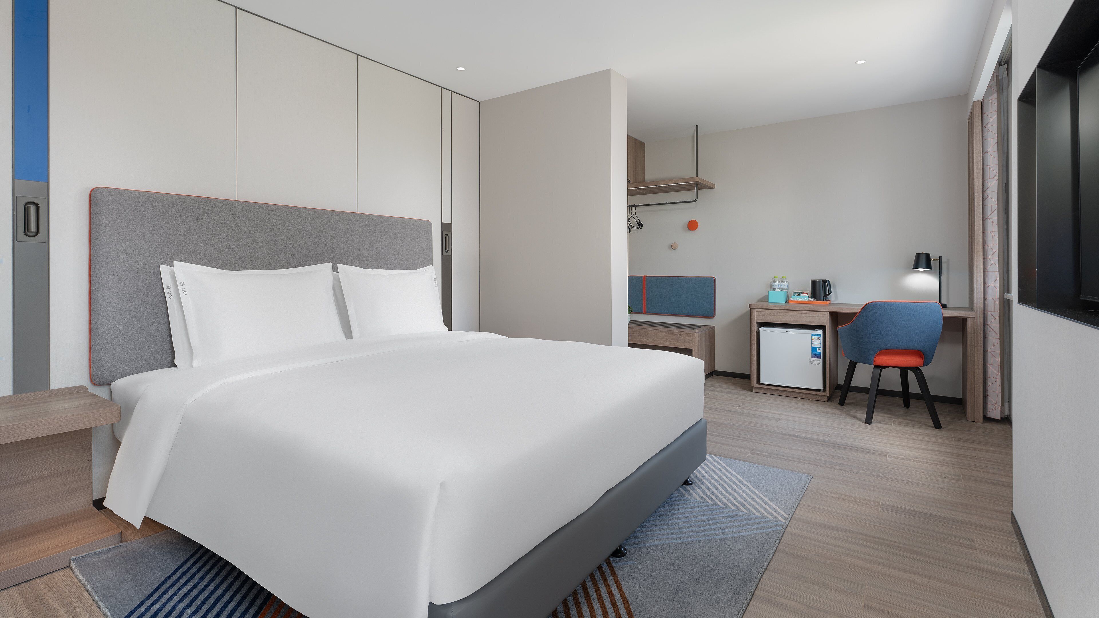 A modern hotel room with a large bed, a desk with a chair, a small refrigerator, and a TV.