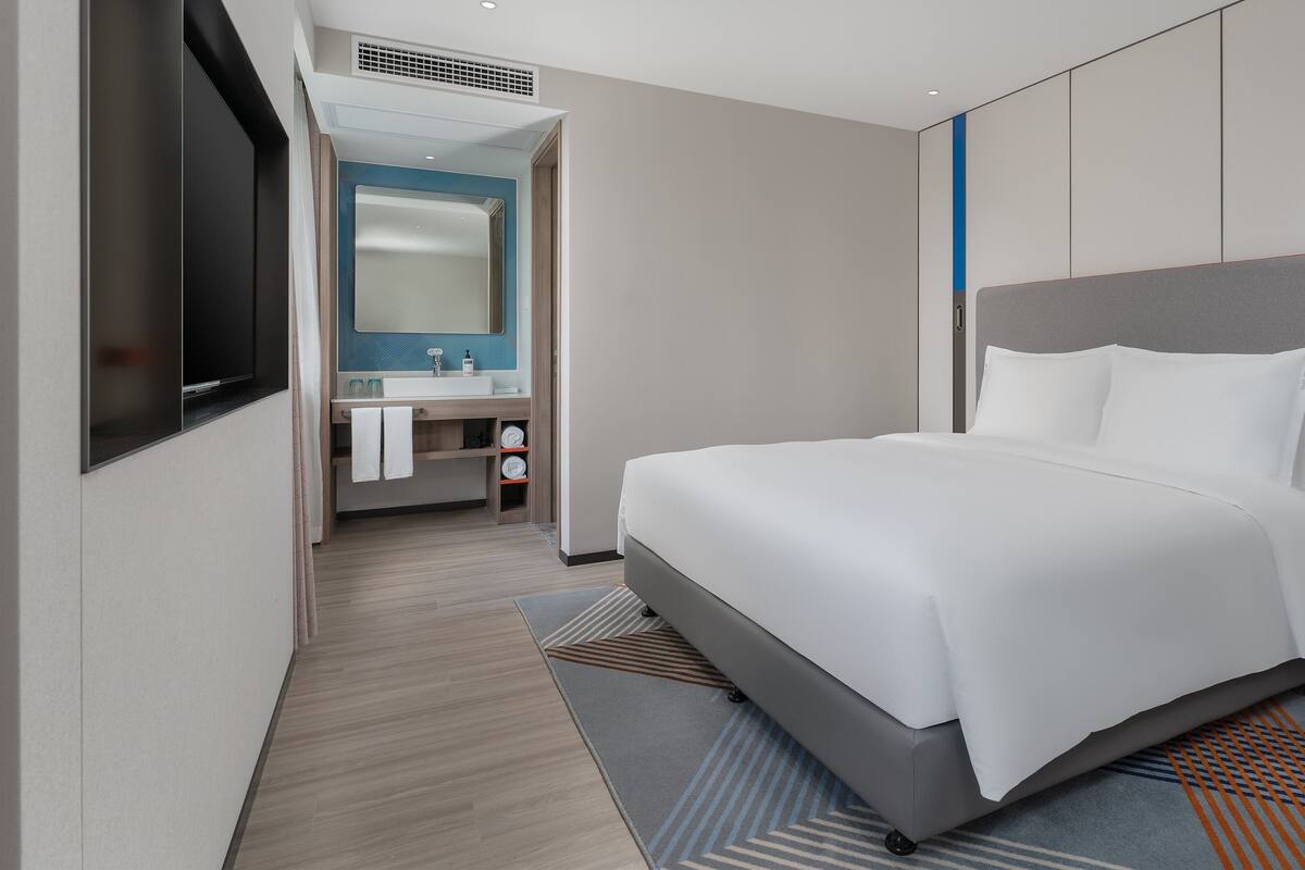 A modern hotel room with a large bed, a built-in TV, a bathroom with a vanity, and a shower.