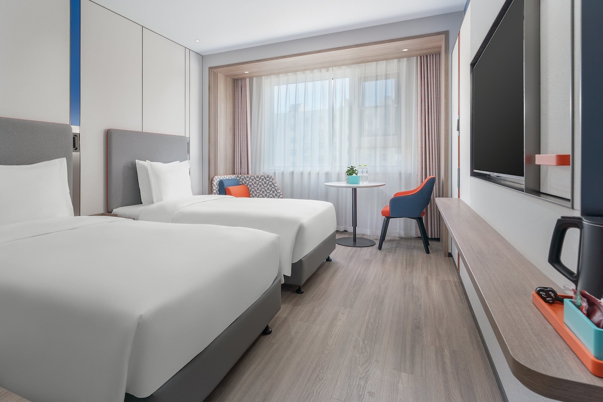 A hotel room with two beds, a desk, a chair, a TV, and a window with curtains.