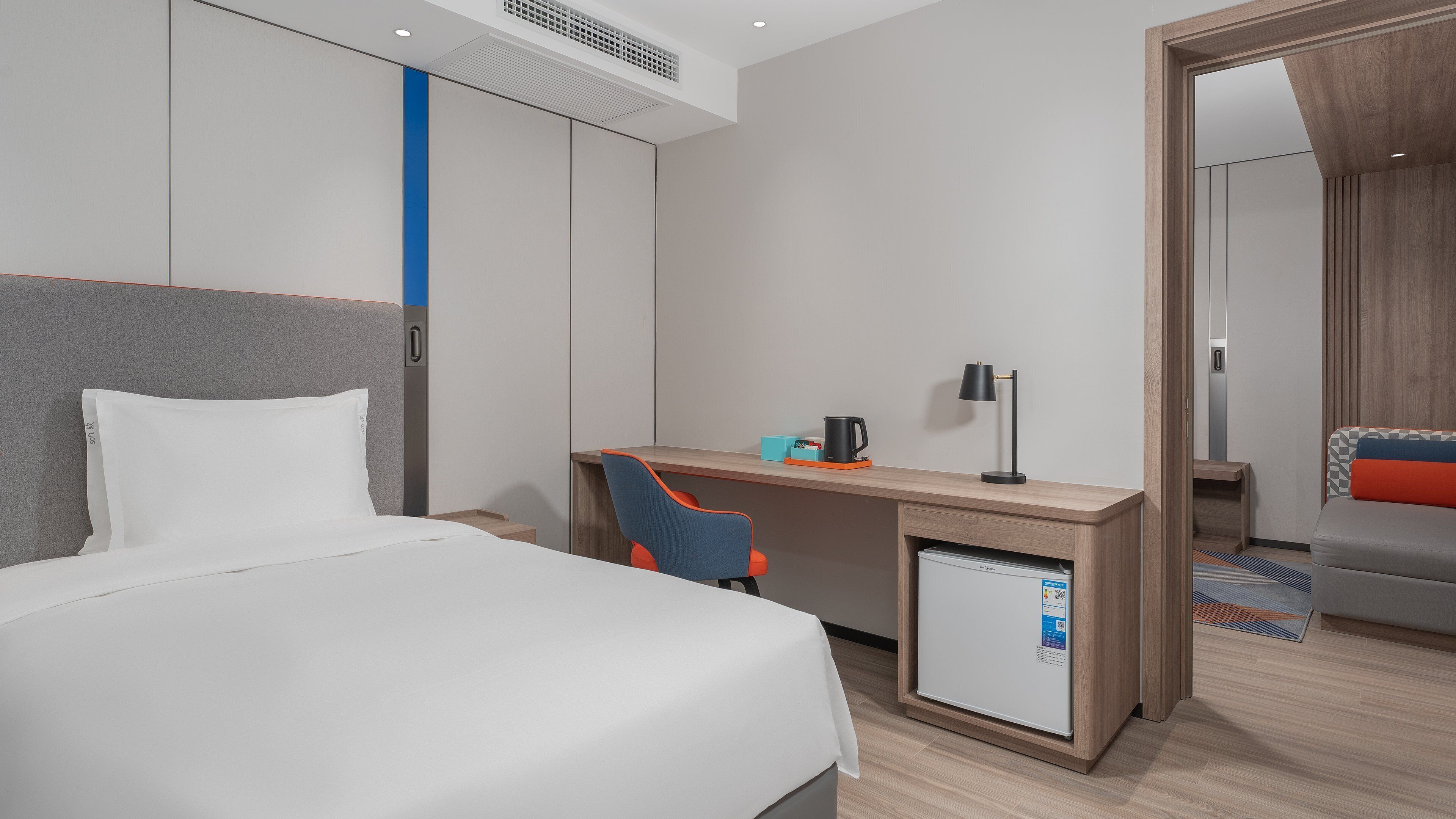 A modern hotel room with a bed, a desk, a chair, a coffee maker, and a mini-fridge.