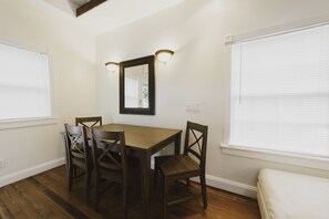 Un coin repas avec une table en bois et quatre chaises, un grand miroir au-dessus de la table et deux fenêtres équipées de stores.