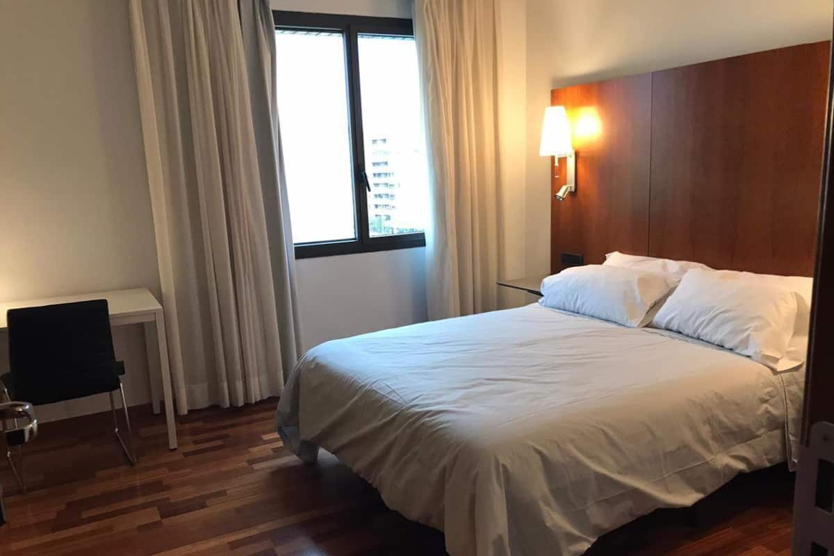 Une chambre d’hôtel avec un plancher en bois, un lit avec du linge de lit blanc, une petite table avec une chaise et une fenêtre avec des rideaux.