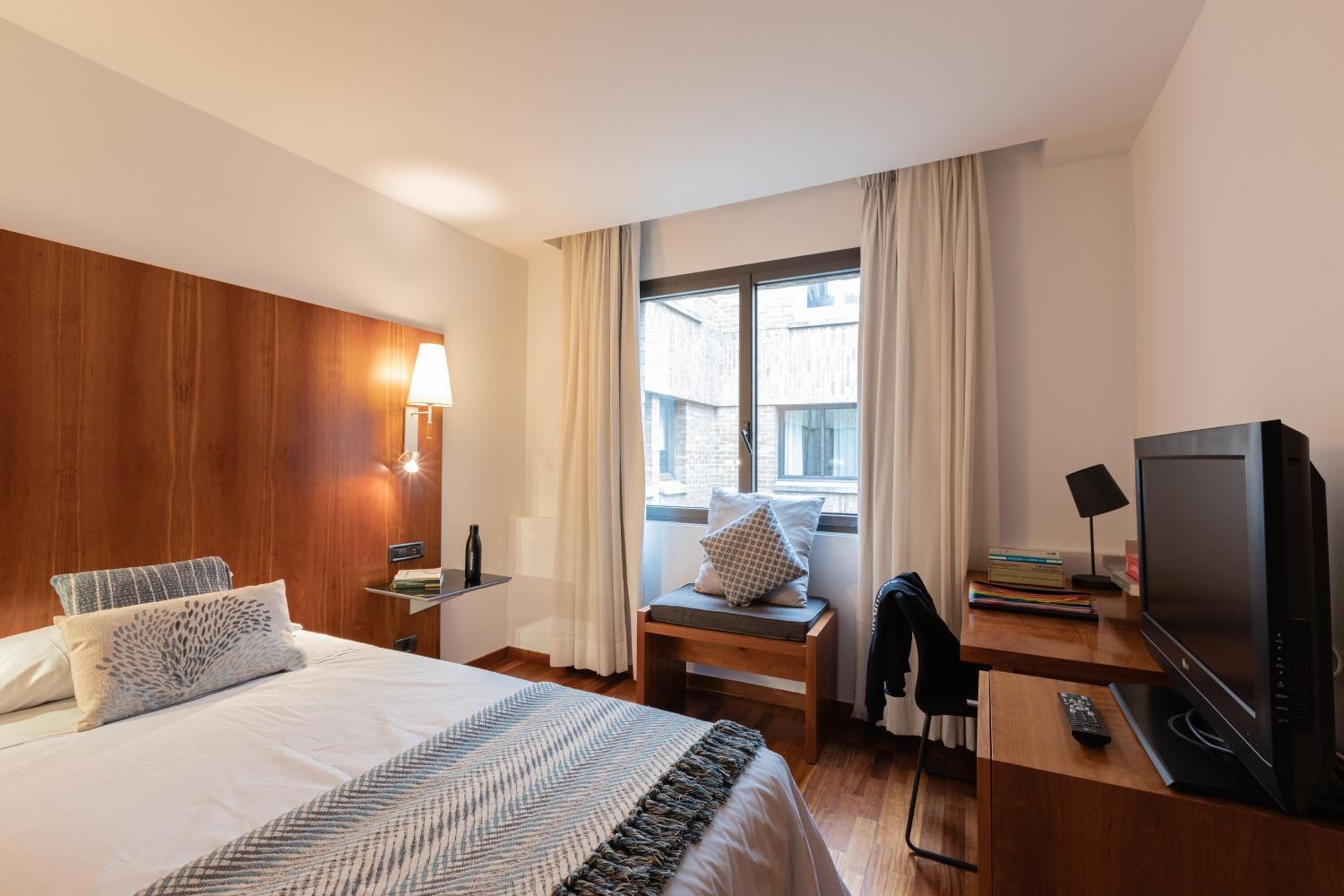 Habitación de hotel con cama, escritorio con televisor, lámpara y ventana con cortinas.