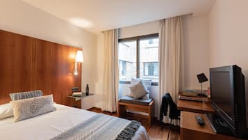 Habitación de hotel con cama, escritorio con televisor, lámpara y ventana con cortinas.
